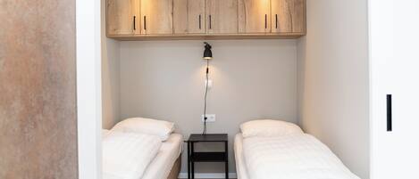 Une petite chambre moderne avec deux lits simples, une armoire en bois et une table de chevet avec une lampe.