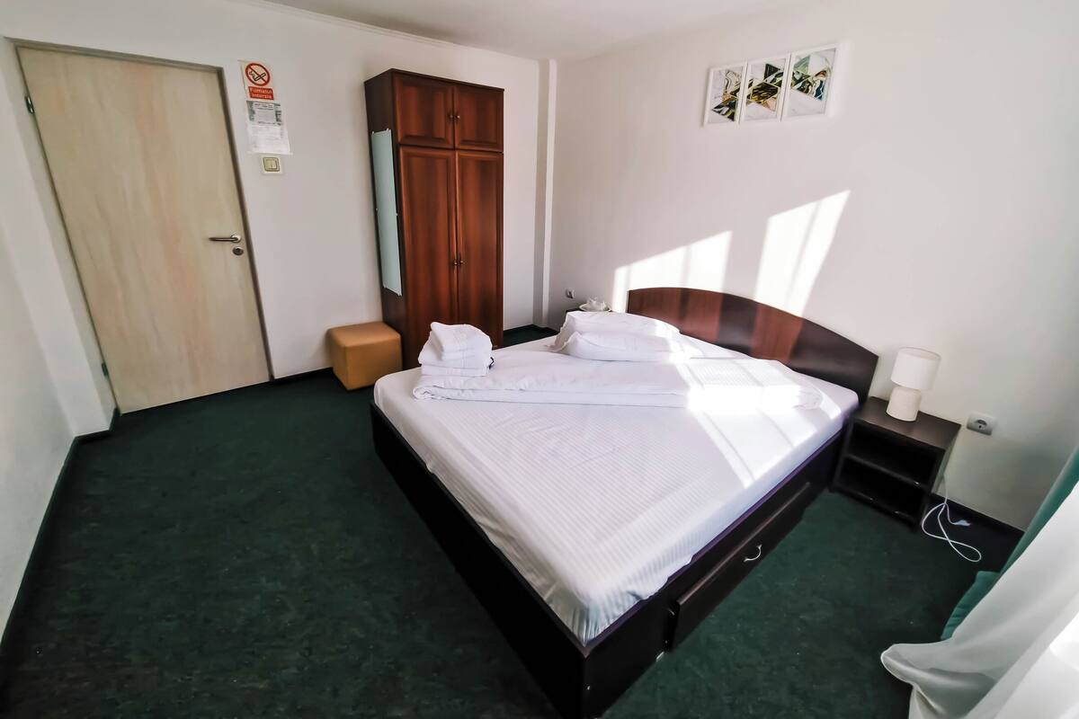 Une chambre d’hôtel comprenant un lit, une armoire en bois, un petit tabouret, une table de chevet avec une lampe et une œuvre d’art accrochée au mur.