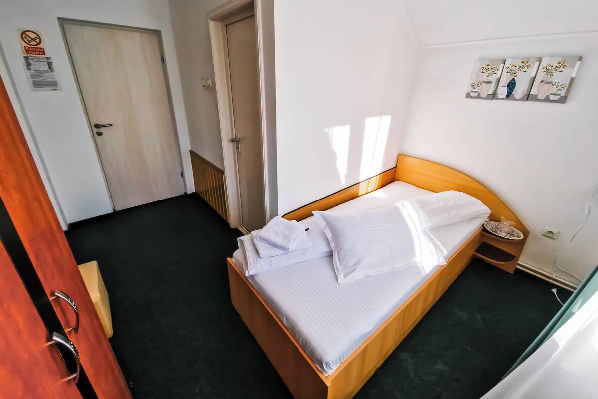 Un lit simple avec une literie blanche, dans une pièce dont le sol est foncé, avec une porte en bois et un interrupteur fixé au mur.