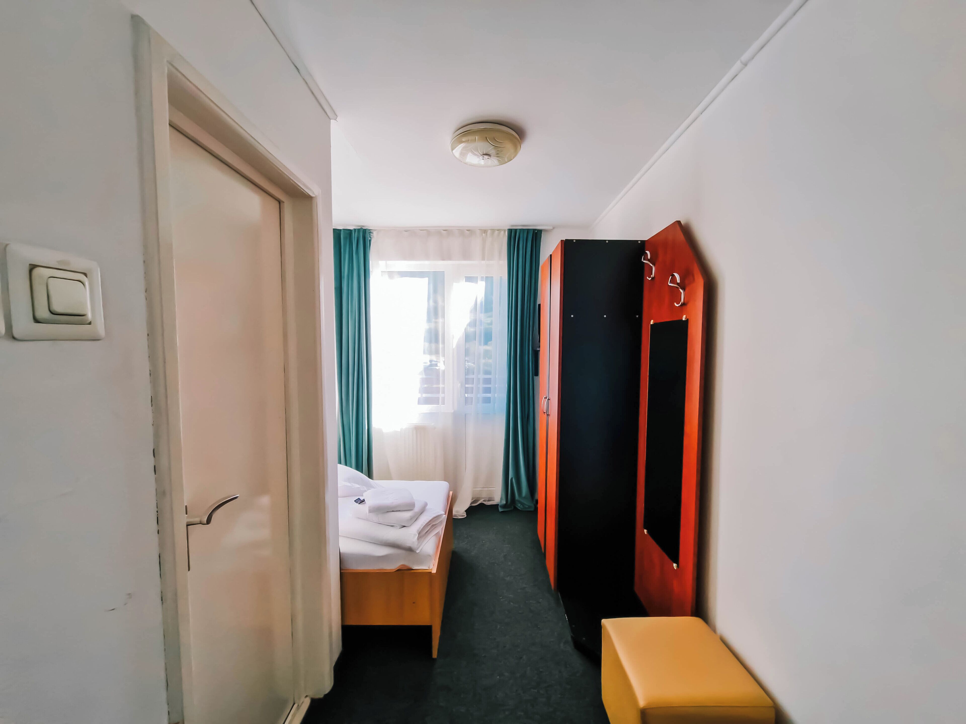 A hotel room with a bed, a bench, a wardrobe, and a window with curtains.