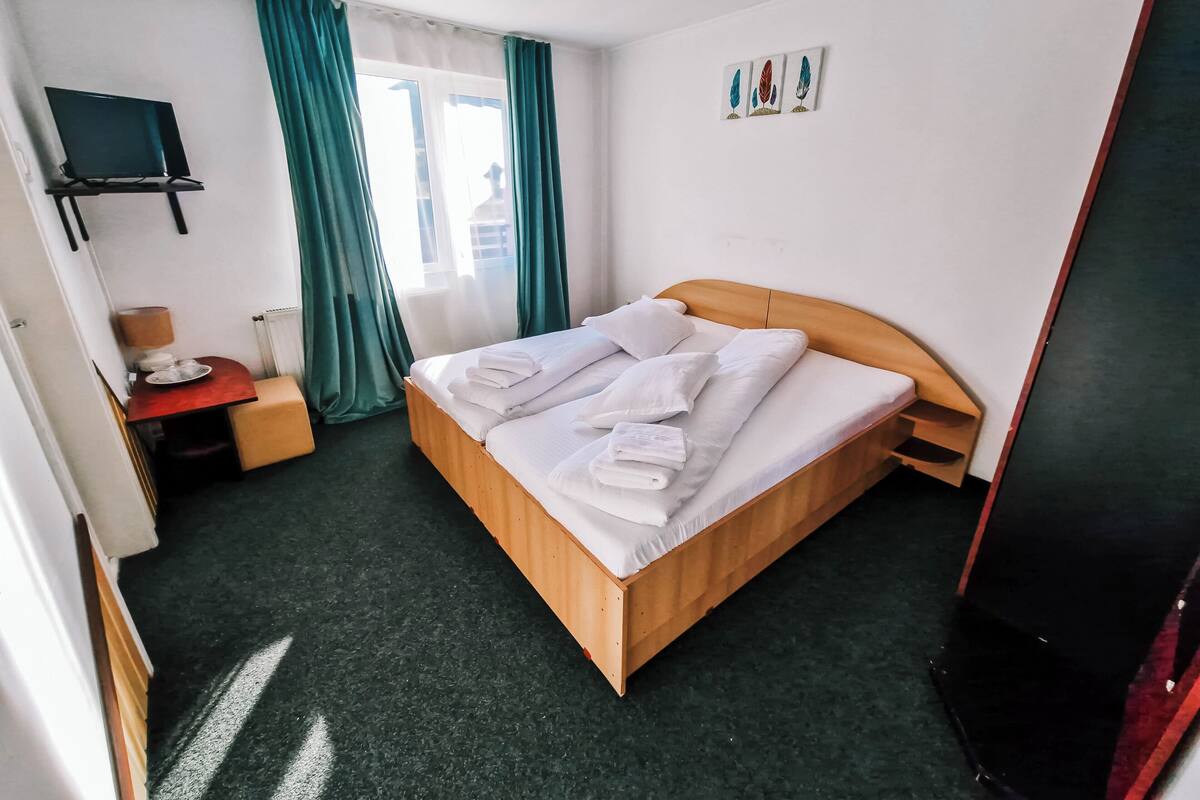 Une chambre d’hôtel avec un lit en bois, un bureau avec une lampe, une télévision et une fenêtre avec des rideaux.