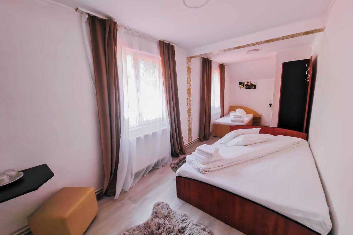 Une chambre d’hôtel avec un lit, une petite table, une chaise, un miroir et une fenêtre avec des rideaux.