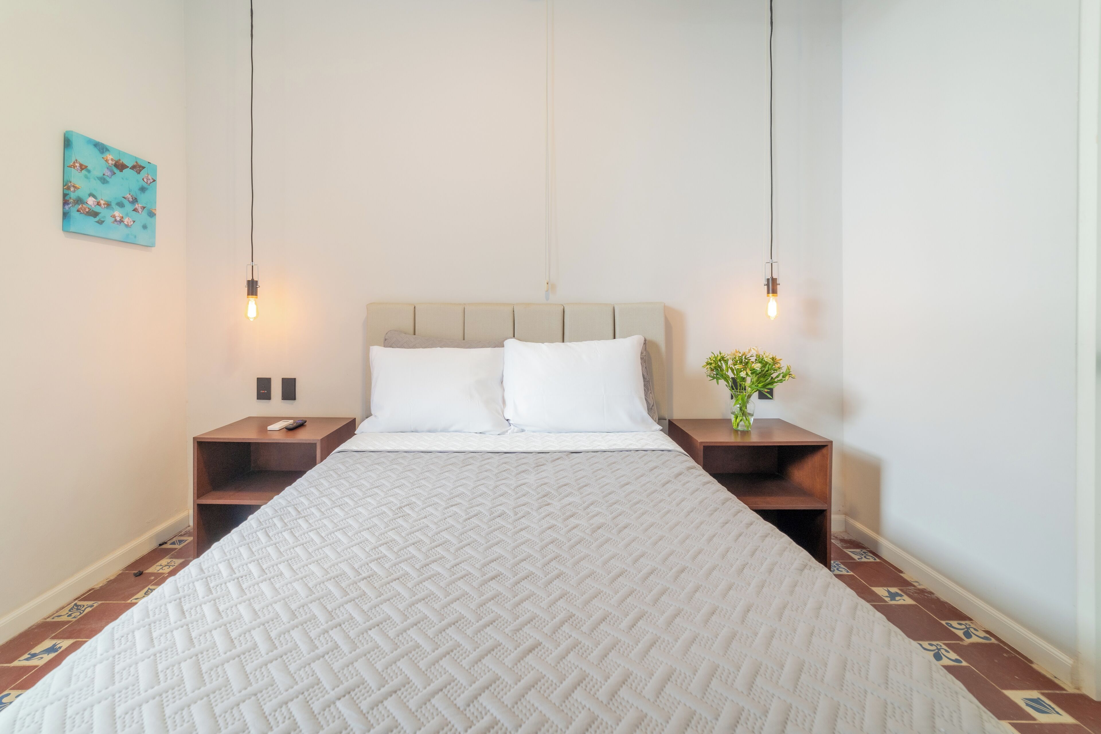 Un lit bien fait, recouvert d’une courtepointe blanche, deux tables de chevet avec des lampes, une petite plante sur l’une des tables et une œuvre d’art bleue accrochée au mur.