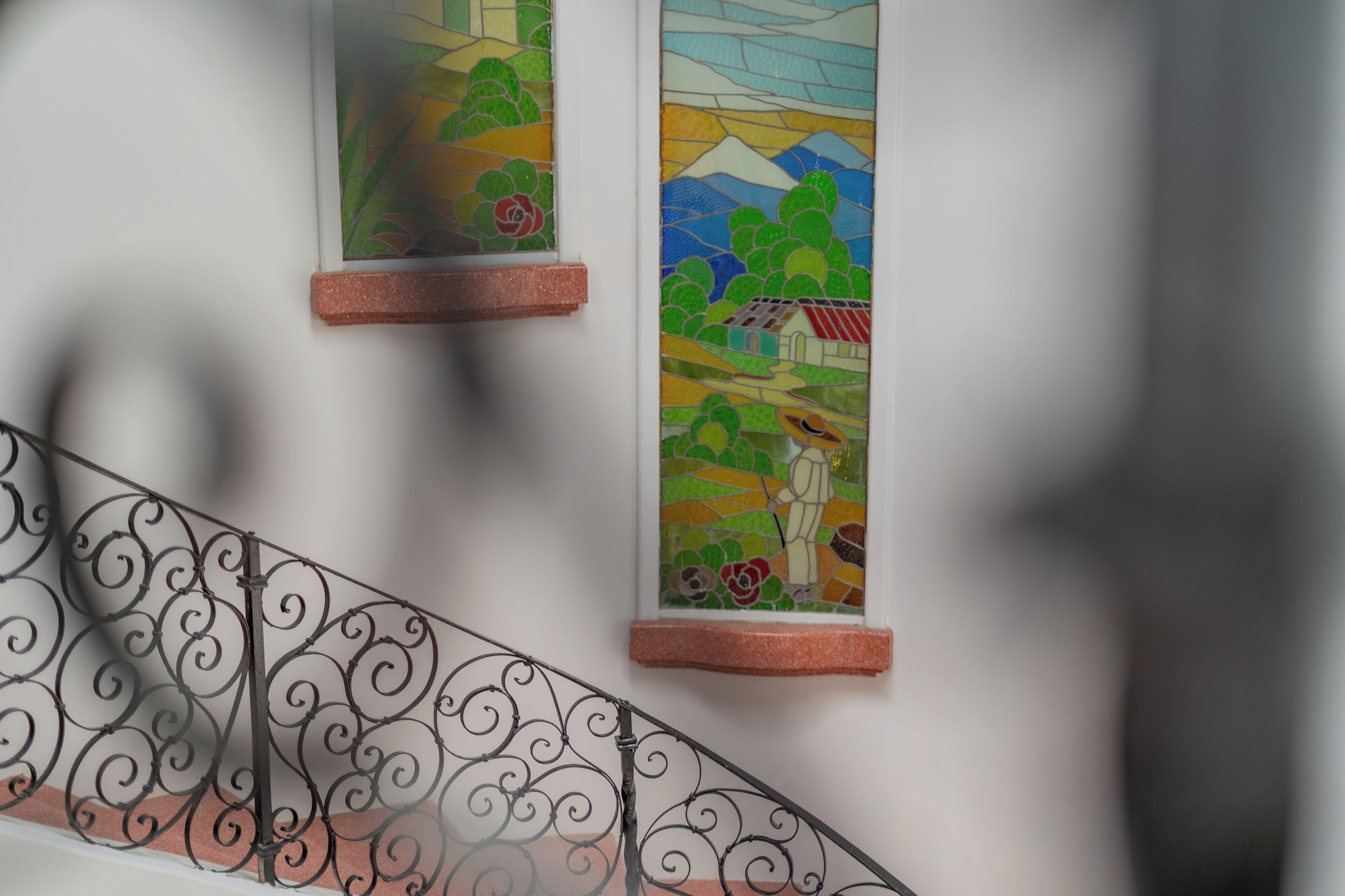 Una escalera con barandilla metálica decorativa y dos vidrieras que representan paisajes.