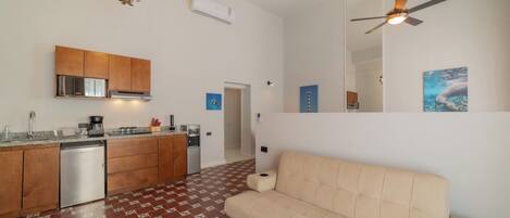 Una cocina moderna con lavavajillas, cafetera y fregadero. En la sala de estar, hay un sofá color beige y un ventilador de techo.