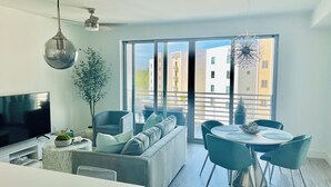 Ein modernes Wohnzimmer mit einer Couch, einem Esstisch und Stühlen. Es gibt einen Fernseher, eine Pflanze und ein großes Fenster mit Blick auf Gebäude.
