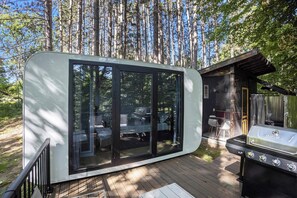 A modern, glass-enclosed cabin with a deck, a barbecue grill, and a patio area. - Hidden Gem Tiny House, Outdoor Screened-In Kitchen (Sears)