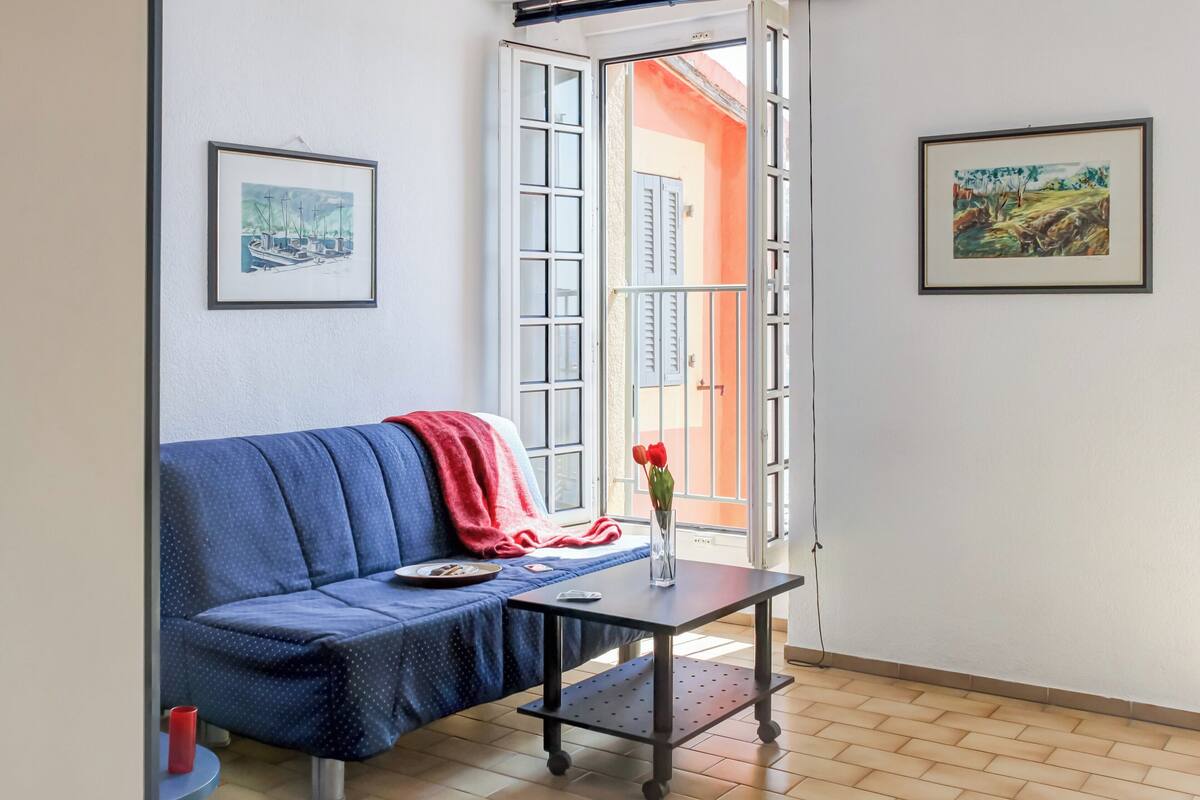 Una habitación con un sofá azul, una pequeña mesa con un jarrón de flores rojas, y dos cuadros en las paredes.