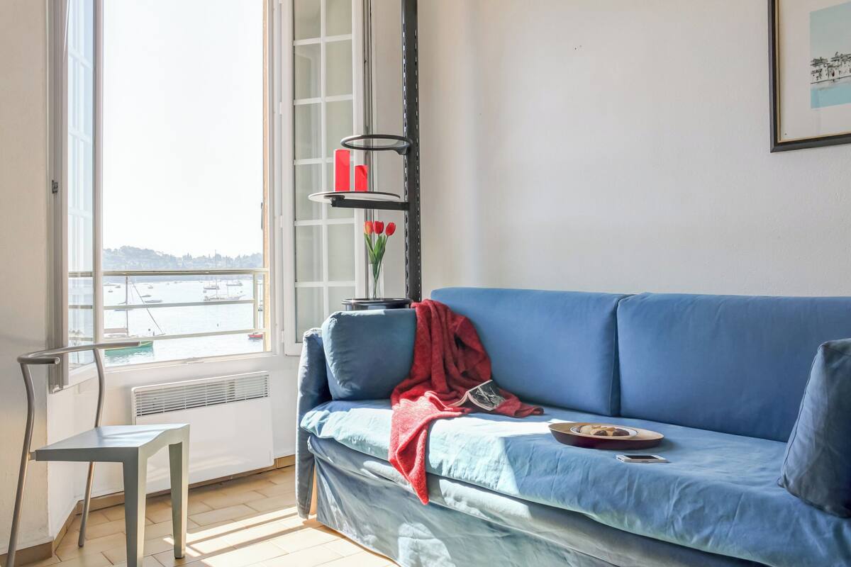 Una habitación con sofá azul, una mesita, un radiador, un ventanal con vista al agua y una estantería con un jarrón de flores rojas.