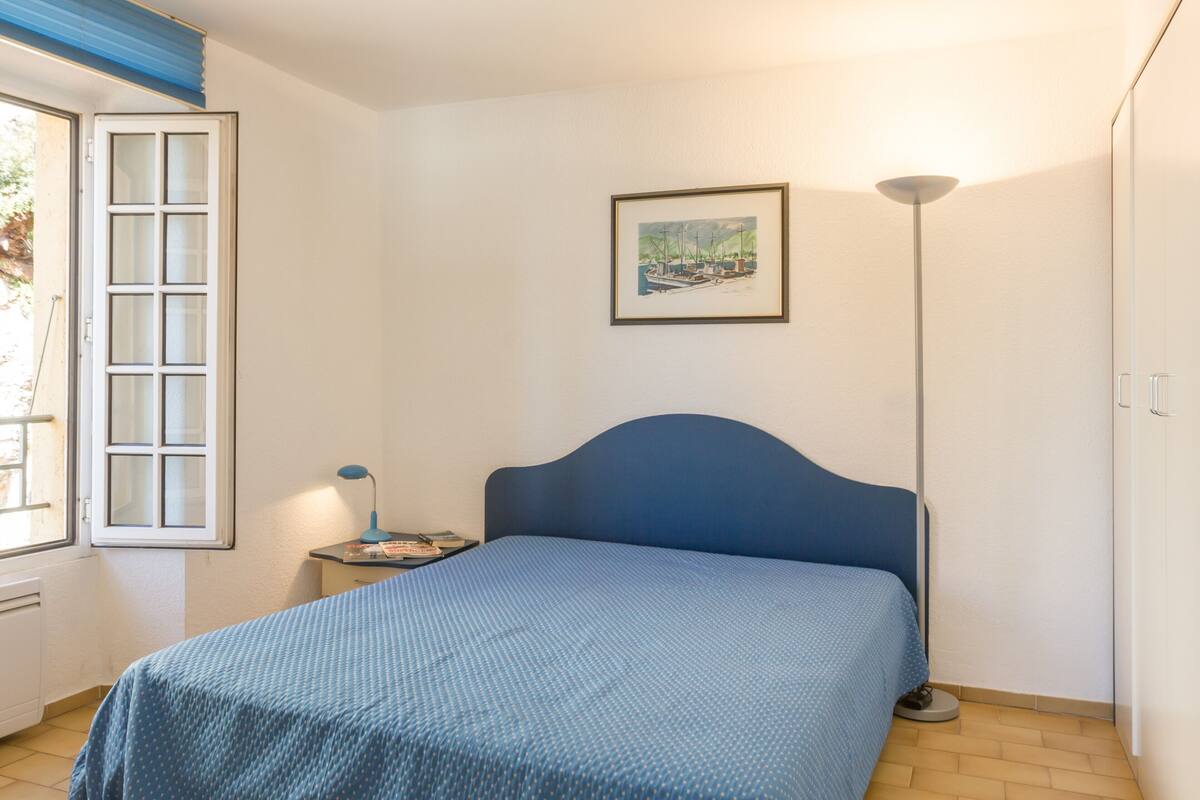 Un dormitorio con cama, mesita de noche, lámpara, ventana con persianas y un cuadro en la pared.