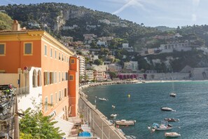 A coastal town with buildings along the waterfront, a clear view of the sea, and a rocky hillside in the background. - Résidence Le Palais de la Marine (Villefranche-sur-Mer)