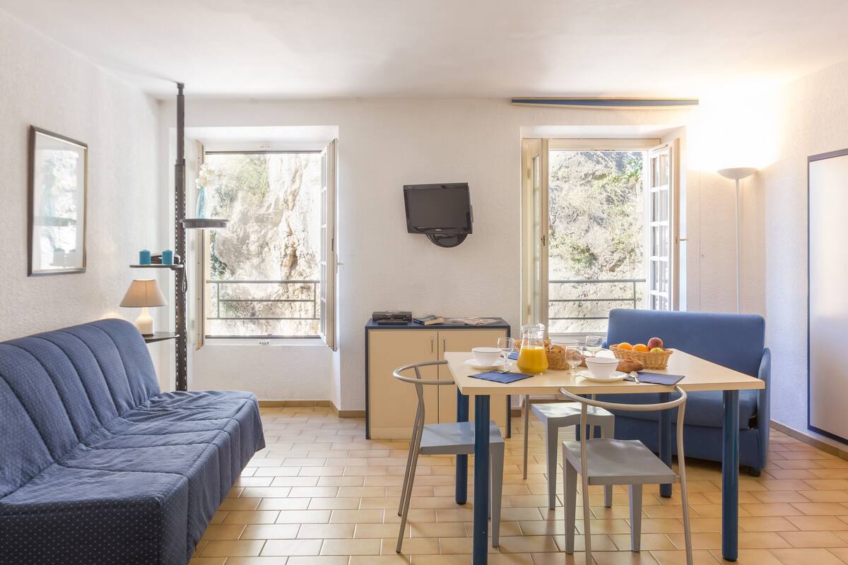 Un espacio habitable compacto con un sofá, una pequeña mesa de comedor con sillas, un televisor montado en la pared y una ventana que ofrece vistas a árboles.