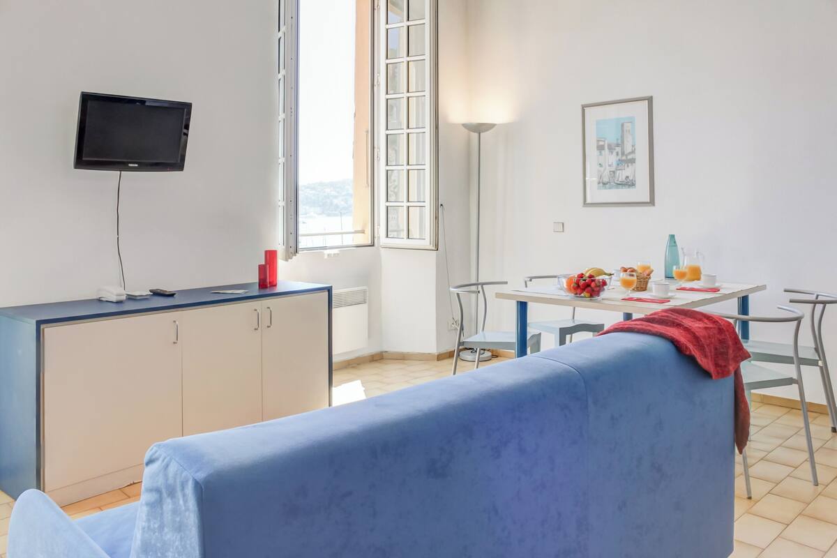 Una habitación con sofá azul, una mesa de comedor con sillas, televisor en la pared y ventana con vistas.