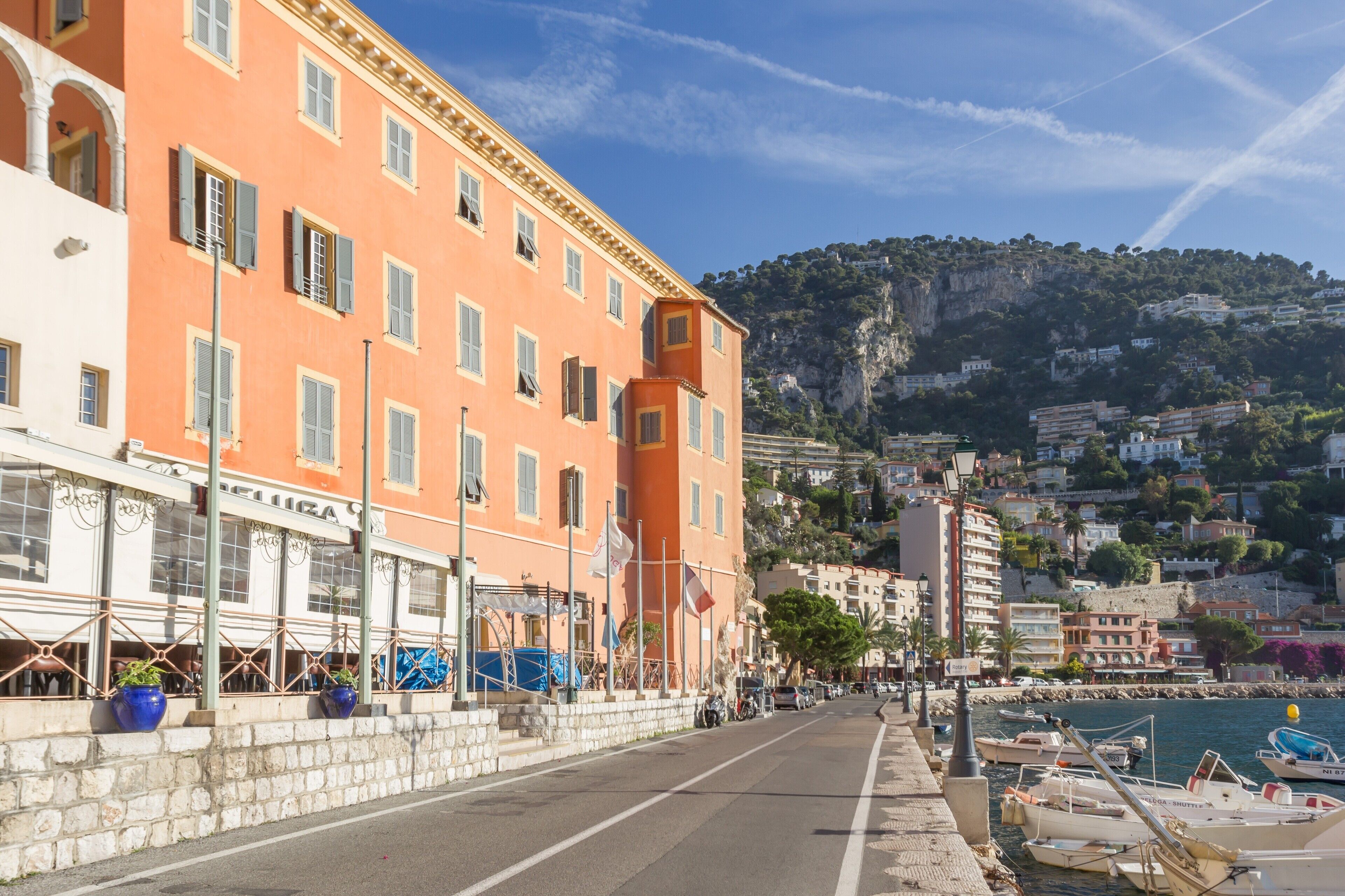 A coastal street with buildings, a clear sky, and boats docked at the harbor.