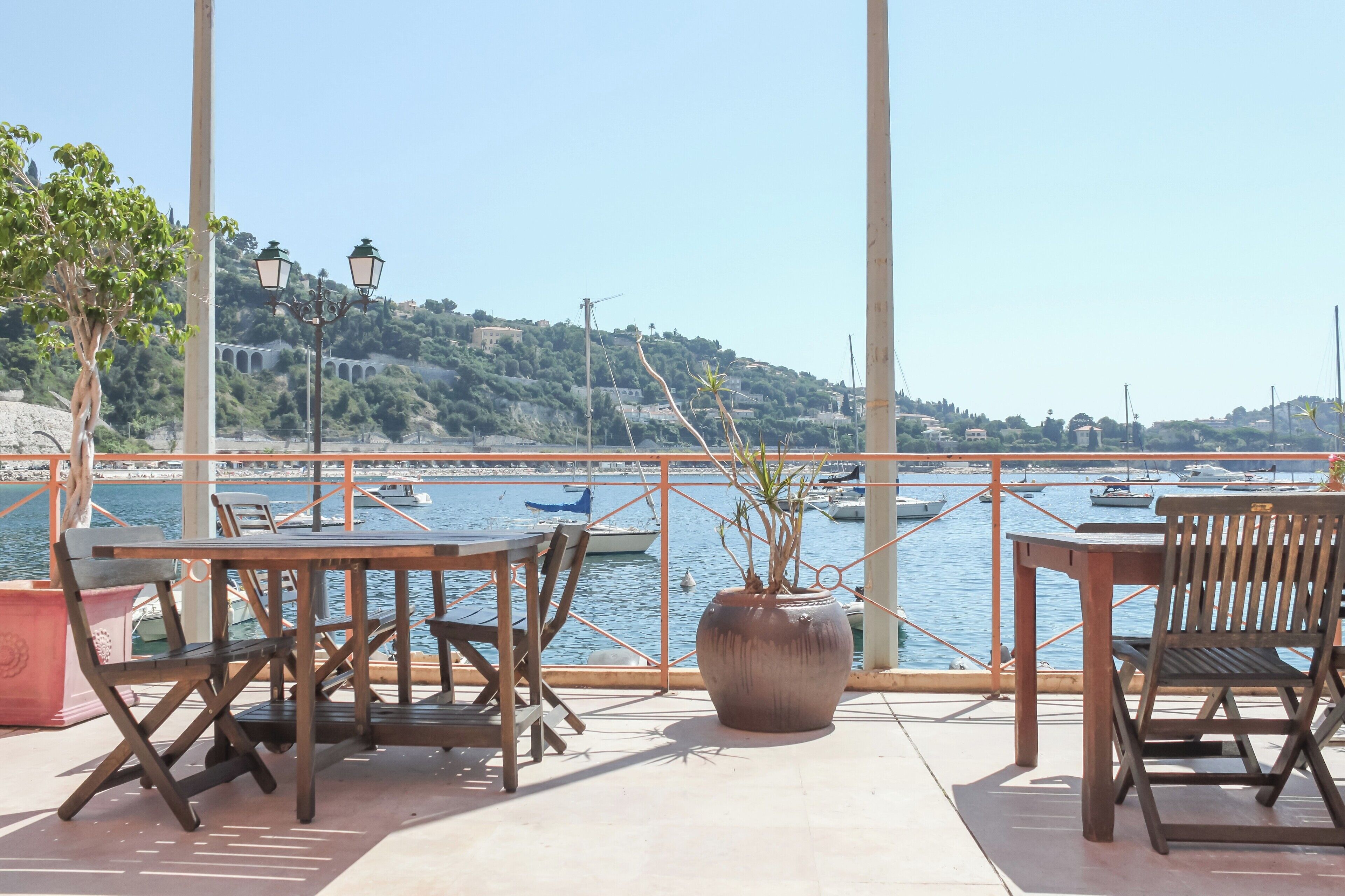 Een terras met tafels en stoelen met uitzicht op een jachthaven vol boten en een heuvellandschap.