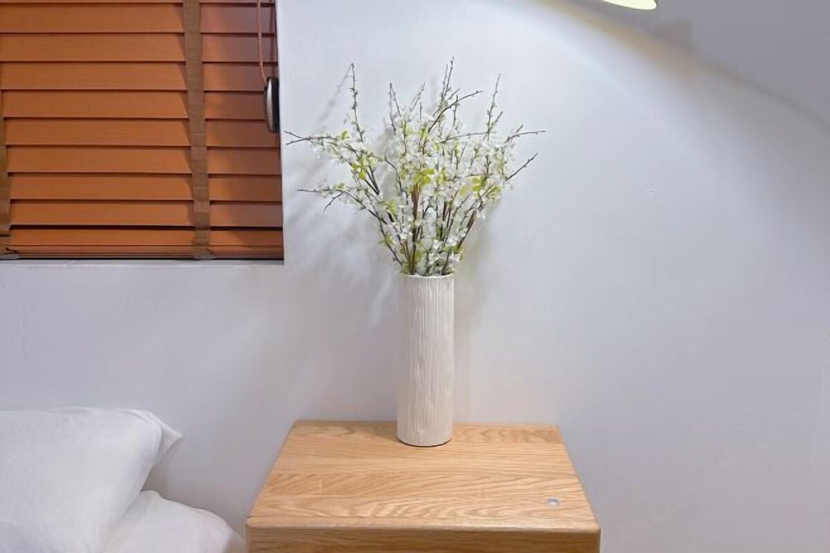 A white vase with branches on a wooden nightstand in a room with a white wall and a wooden window blind.