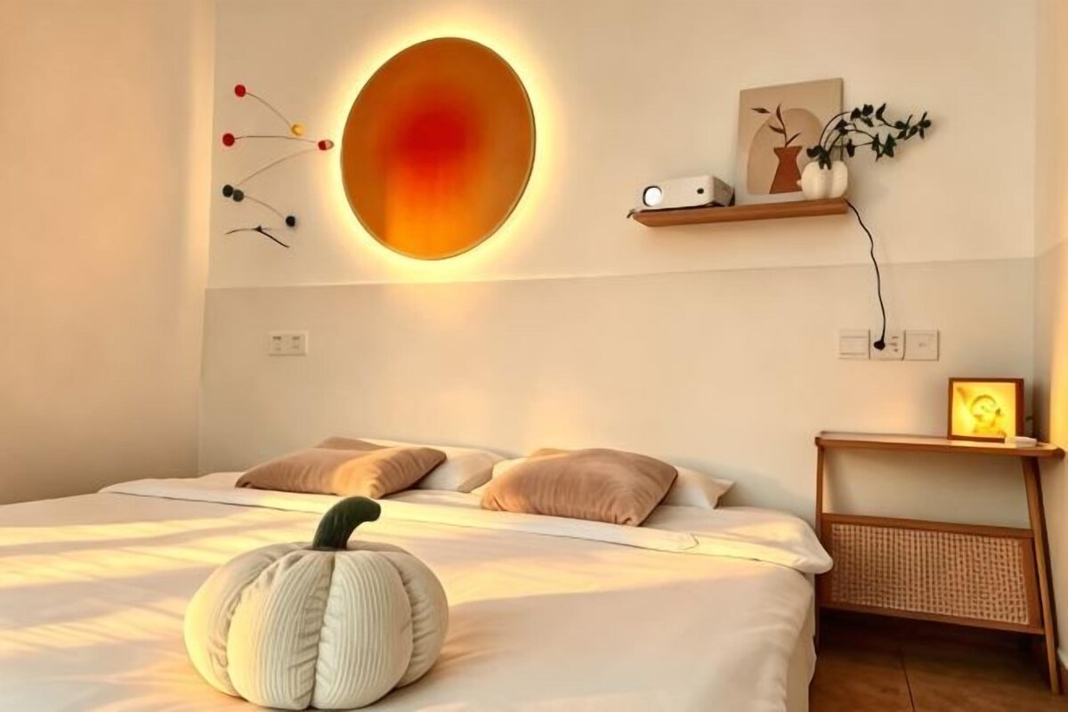 A bedroom with a bed, pillows, a wall-mounted light, a bedside table, and a wall decoration.