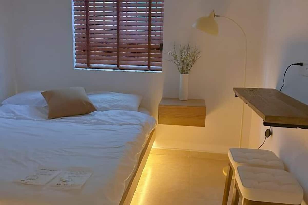 A bedroom with a bed, a bedside table with a lamp, a small bedside plant, and a wooden shelf.