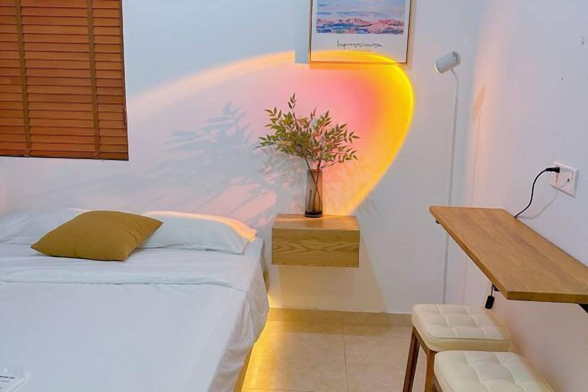 A bedroom with a bed, a bedside table with a plant, a bedside lamp, and a wooden bench.