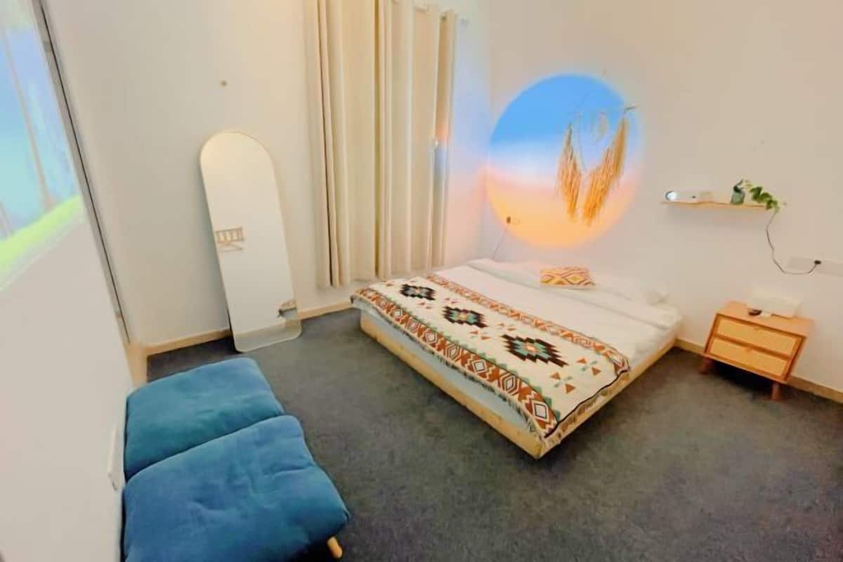 A bedroom with a bed, a blue chair, a bedside table, a wall-mounted light, and a shelf with plants.