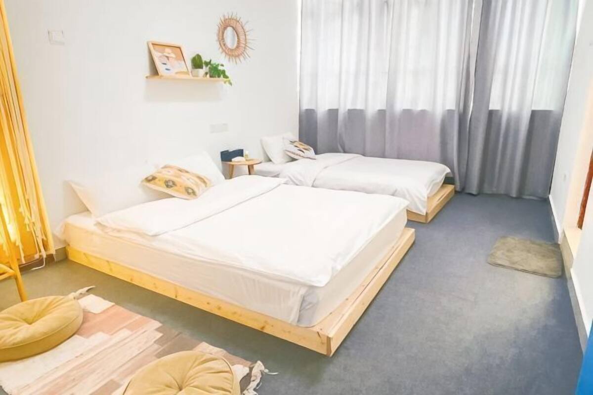 A room with two beds, a small table, a wall-mounted shelf, and a window with curtains.
