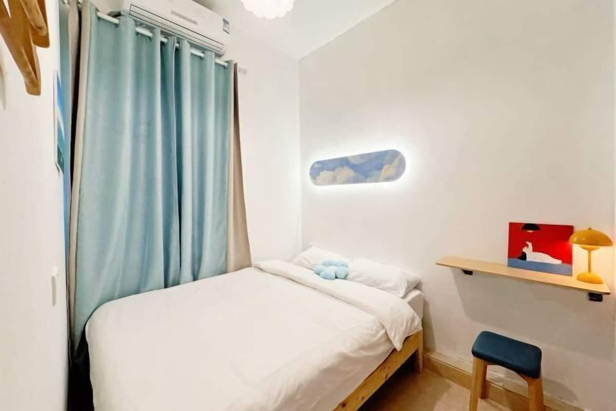 A bedroom with a bed, a desk, a chair, a shelf with a lamp, and a wall-mounted light fixture.