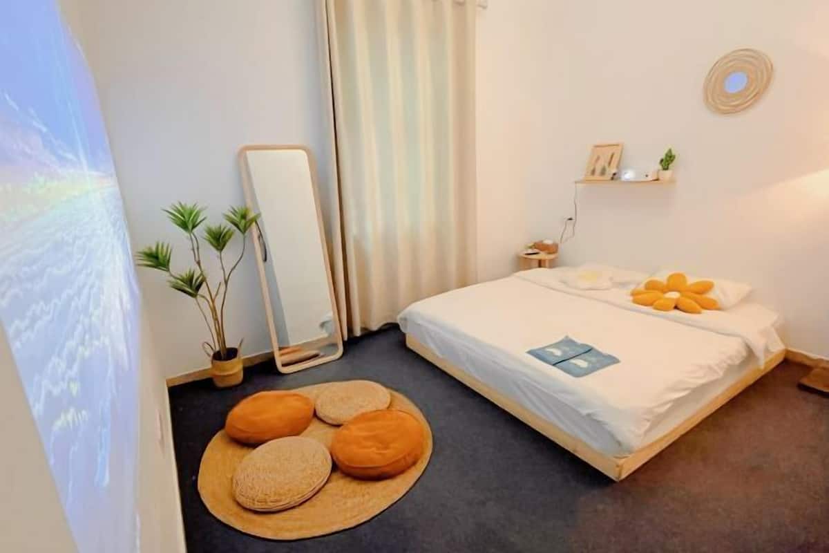 A bedroom with a bed, a mirror, a bedside table, a wall-mounted shelf, and a potted plant.