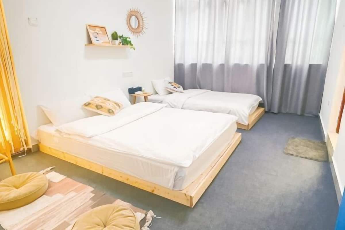 A room with two beds, a small table, a wall-mounted shelf, and a window with curtains.