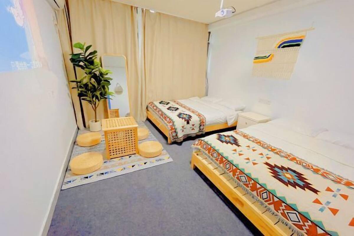 A hotel room with two beds, a small seating area with cushions, a plant, and a wall-mounted shelf.