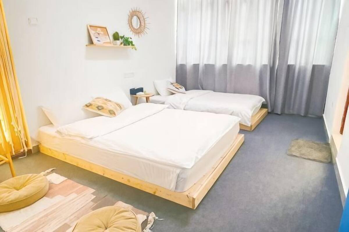 A room with two beds, a small table, a wall-mounted shelf, and a window with curtains.