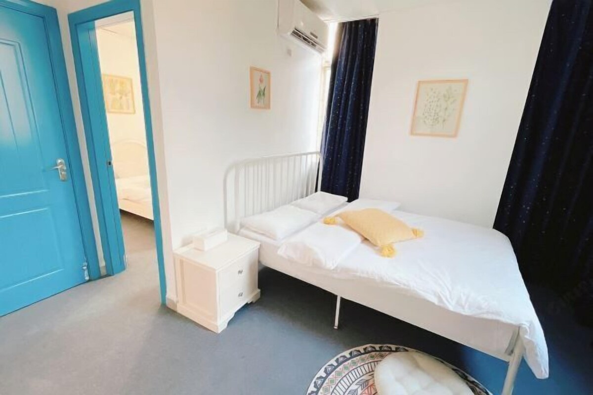 A single-bed room with a white bed, a small bedside table, a white nightstand, a blue door, and a round rug.