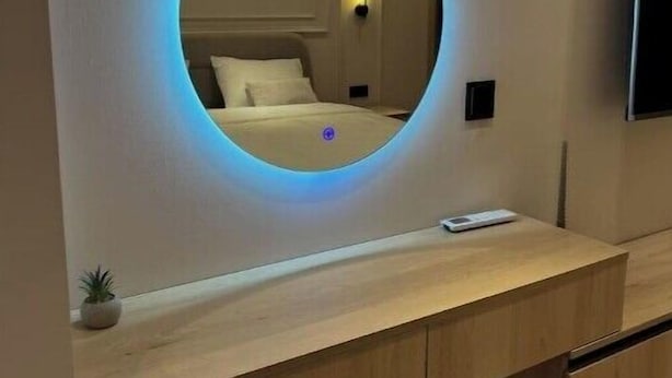 A modern hotel room with a circular mirror on the wall, a bed with white linens, a bedside table with a remote control, and a small potted plant. - Luxurious Central Baku Apartment (Baku)