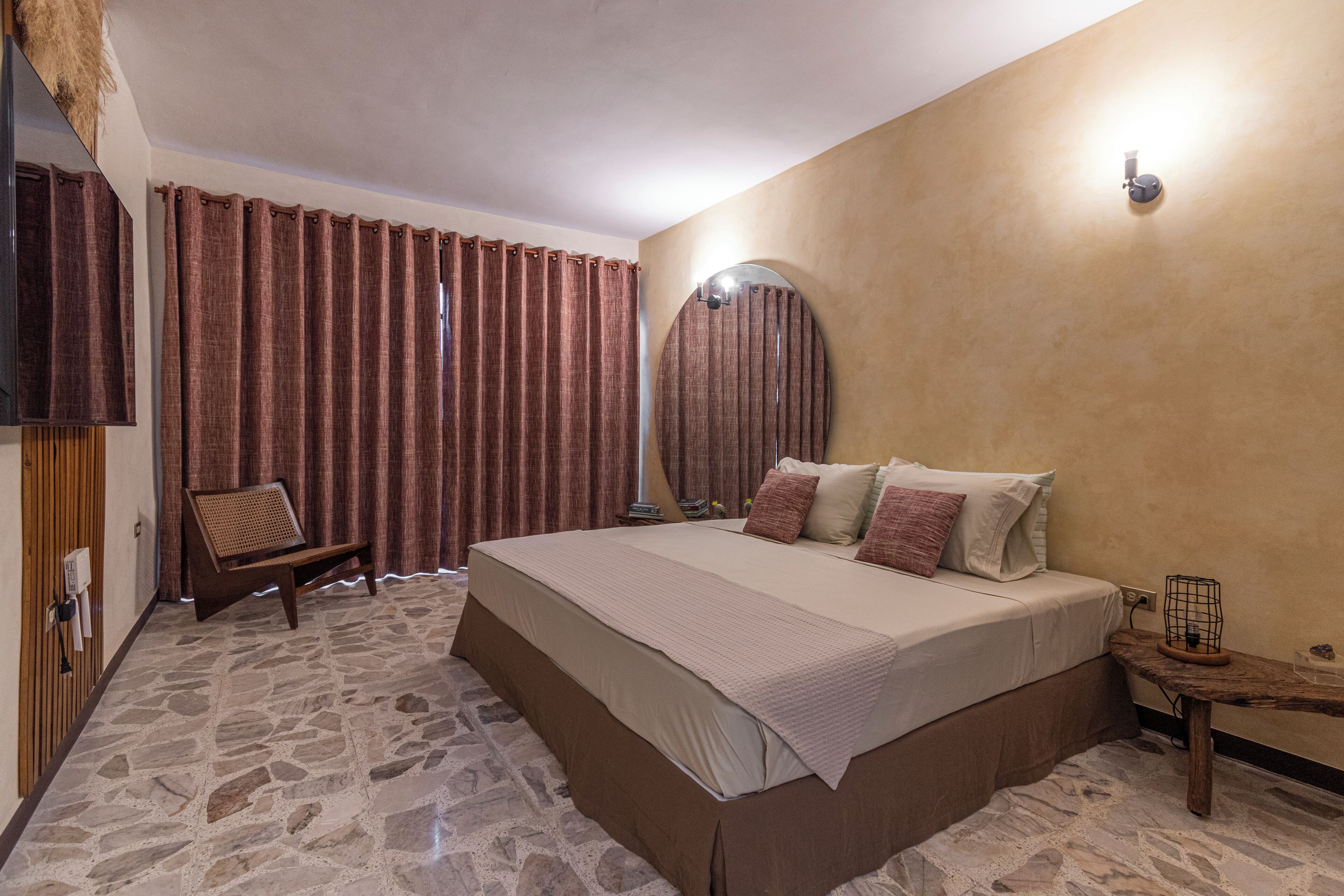 A bedroom with a large bed, a chair, a bedside table, a mirror, and curtains.