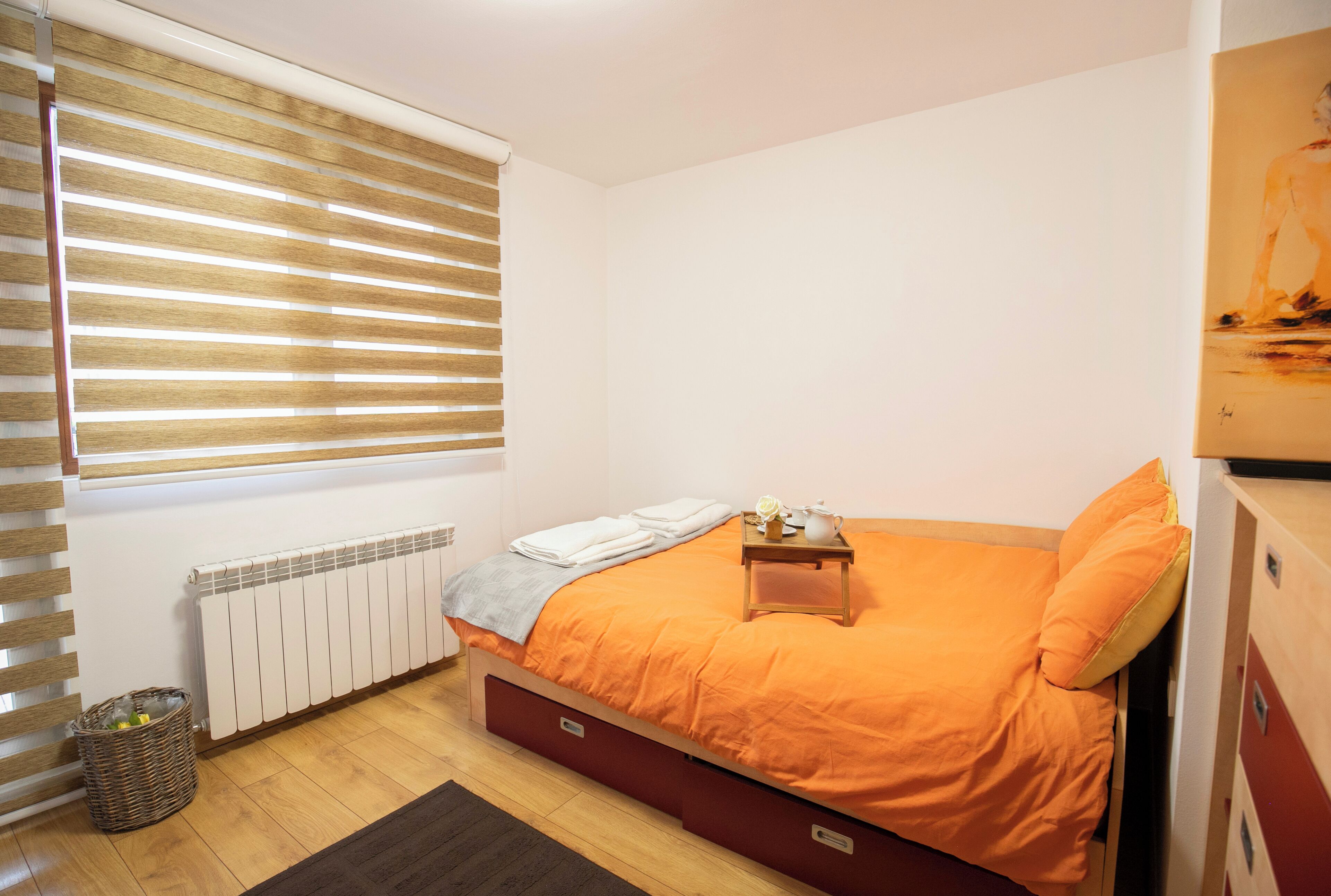 Un dormitorio con cama, mesita de noche, radiador, ventana con persianas, cesta de mimbre y suelo de madera.