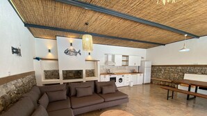 Un espace de vie moderne comprenant un canapé d’angle, une table basse ronde en bois, une cuisine avec des meubles blancs et un mur d’accent en pierre.