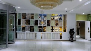 Un hall d’hôtel moderne avec un ascenseur en verre, un mur décoratif orné de panneaux à motifs et un grand lustre.
