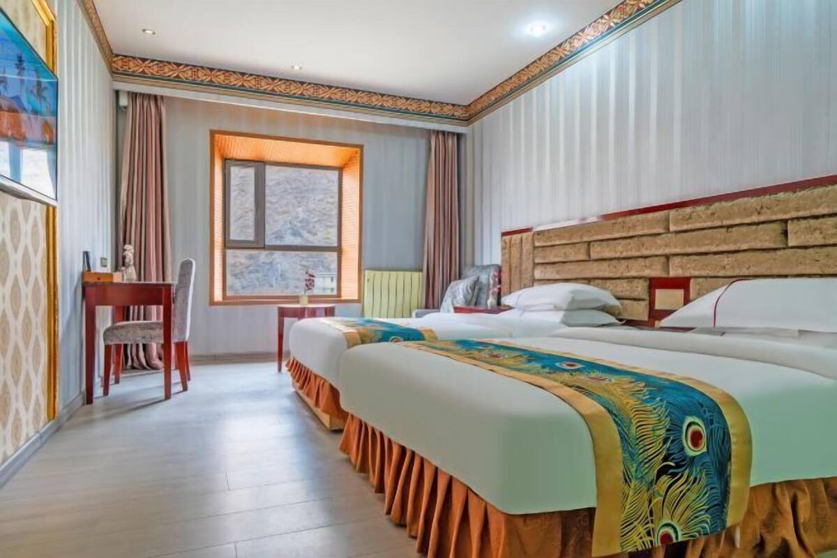 Habitación de hotel con dos camas, cabecera de madera, ventana con cortinas, escritorio con una silla y decoración en la pared.