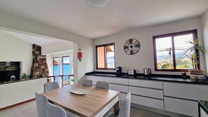 Une cuisine moderne avec une table à manger en bois, des chaises grises, une horloge et une vue sur la mer grâce à de grandes fenêtres.