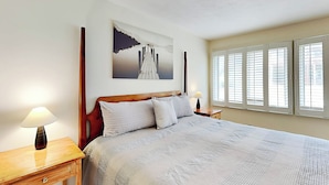 A neatly arranged bedroom with a large bed, two bedside tables, a nightstand, a painting on the wall, and a window with blinds. - Deep Creek Village #33 by Taylor Made Deep Creek (McHenry)