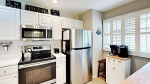 A modern kitchen with white cabinetry, stainless steel appliances, a built-in oven, microwave, and refrigerator. There is a small wooden stool and a coffee maker on the countertop. - Deep Creek Village #33 by Taylor Made Deep Creek (McHenry)