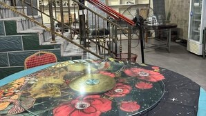 Ein Essbereich mit einem runden Tisch, der mit einer blumigen Tischdecke bedeckt ist, einer Treppe mit roter Handlauf und einem Deckenventilator.