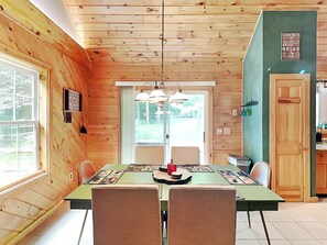 A cozy cabin interior with a wooden ceiling, a dining table with chairs, a kitchen area, and a large window. - Wander Through the Woods by Taylor Made Deep Creek (McHenry)