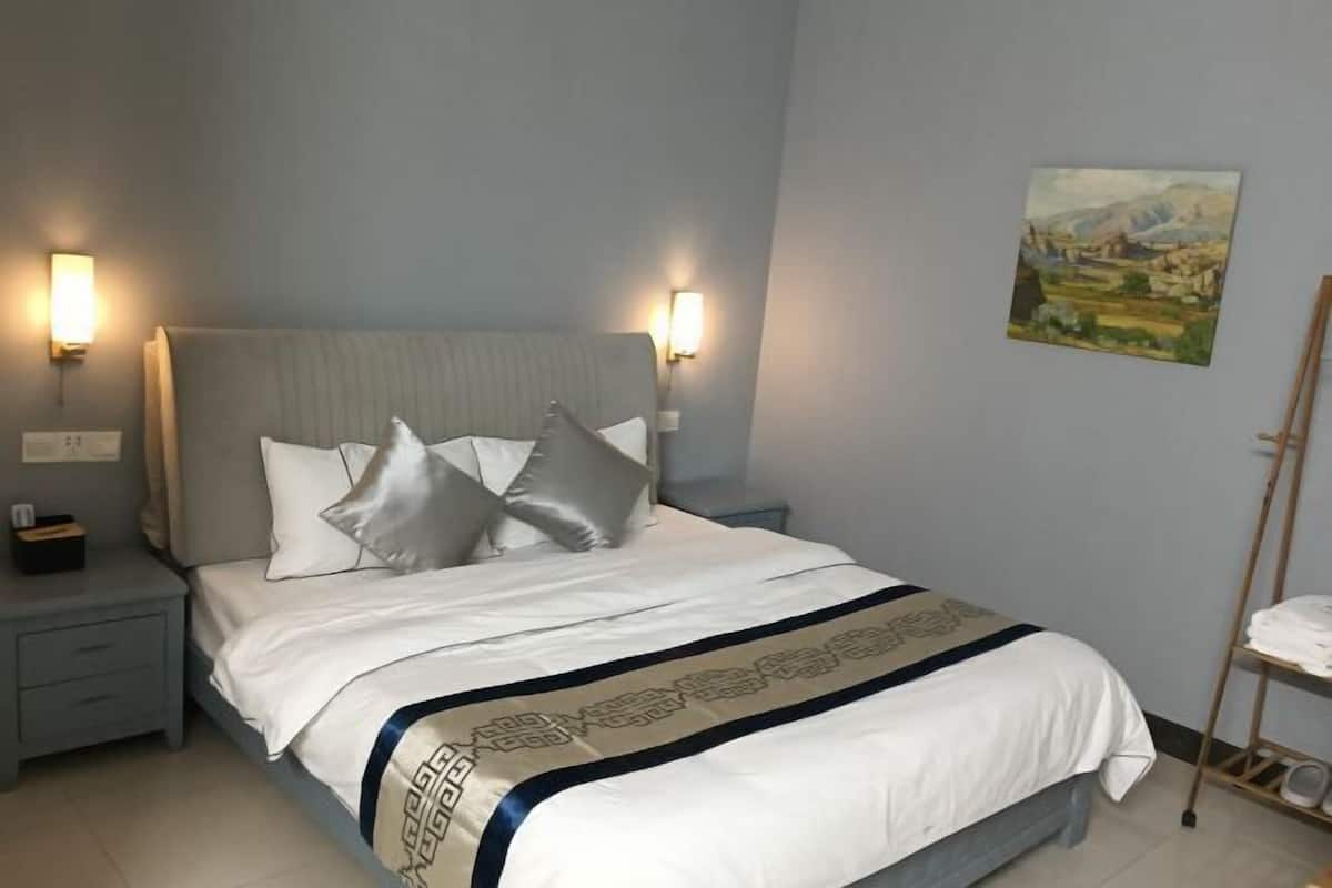 A neatly made bed with a white and beige bedspread, two bedside lamps, a nightstand, and a painting on the wall.