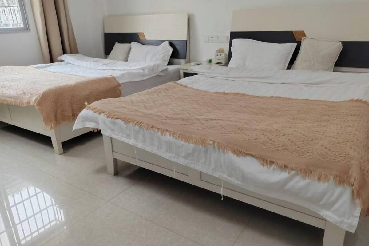 A hotel room with two beds, each with white linens and a beige throw blanket. There are bedside tables with lamps and a small decorative item on one table.