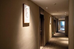 A hotel hallway with a framed artwork on the wall, a door on the right, and a long corridor leading to a bright area at the end.