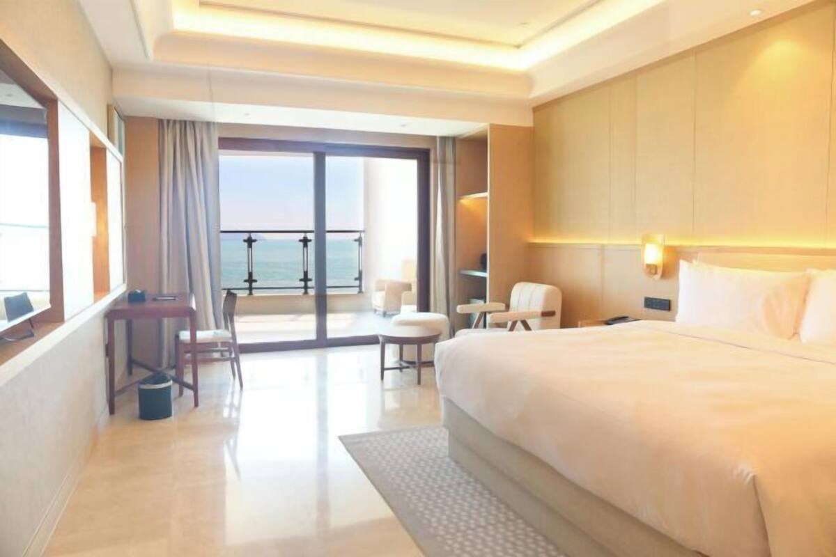 A hotel room with a large bed, a desk, a chair, a small table, a lamp, and a view of the sea.