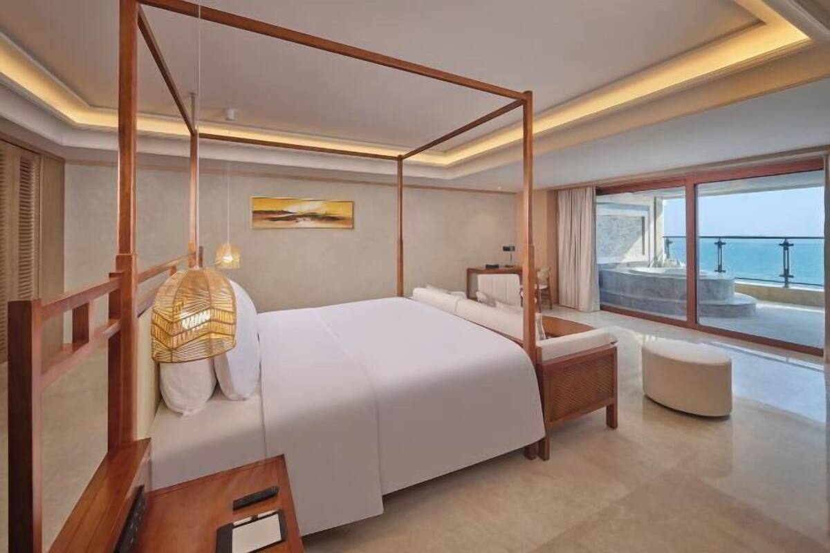 A modern bedroom with a four-poster bed, a bedside lamp, and a view of the sea through large windows.