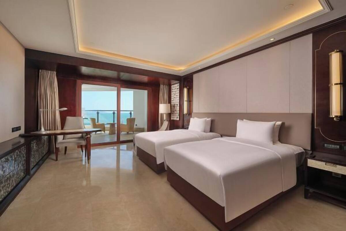 A hotel room with two beds, a desk, a chair, and a view of the sea.