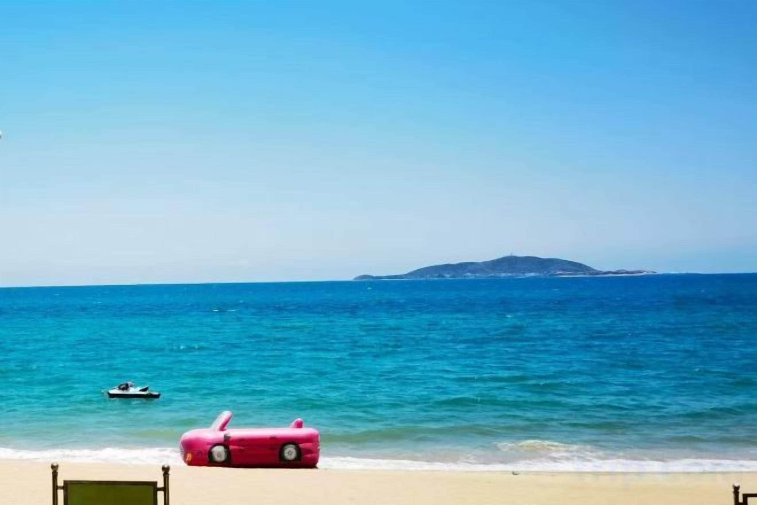 Une plage avec une voiture rouge garée sur le sable, un bateau sur l’eau et une île au loin.