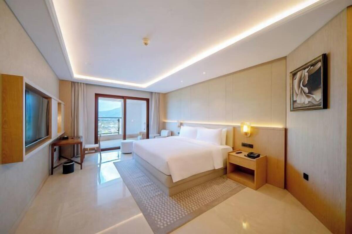A modern hotel room with a large bed, bedside tables, a desk, a chair, a window with curtains, and a painting on the wall.